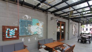 An outdoor cafe with wooden tables and chairs, a concrete floor, and a wall with artwork and a door.
