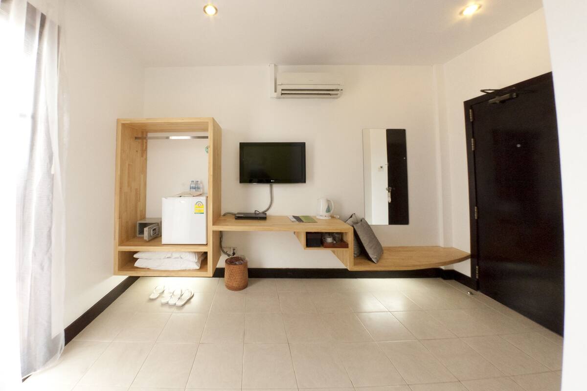 Una habitación de hotel moderna con un armario de madera, un televisor de pantalla plana, un escritorio con un ordenador, una puerta negra y un suelo de baldosas.