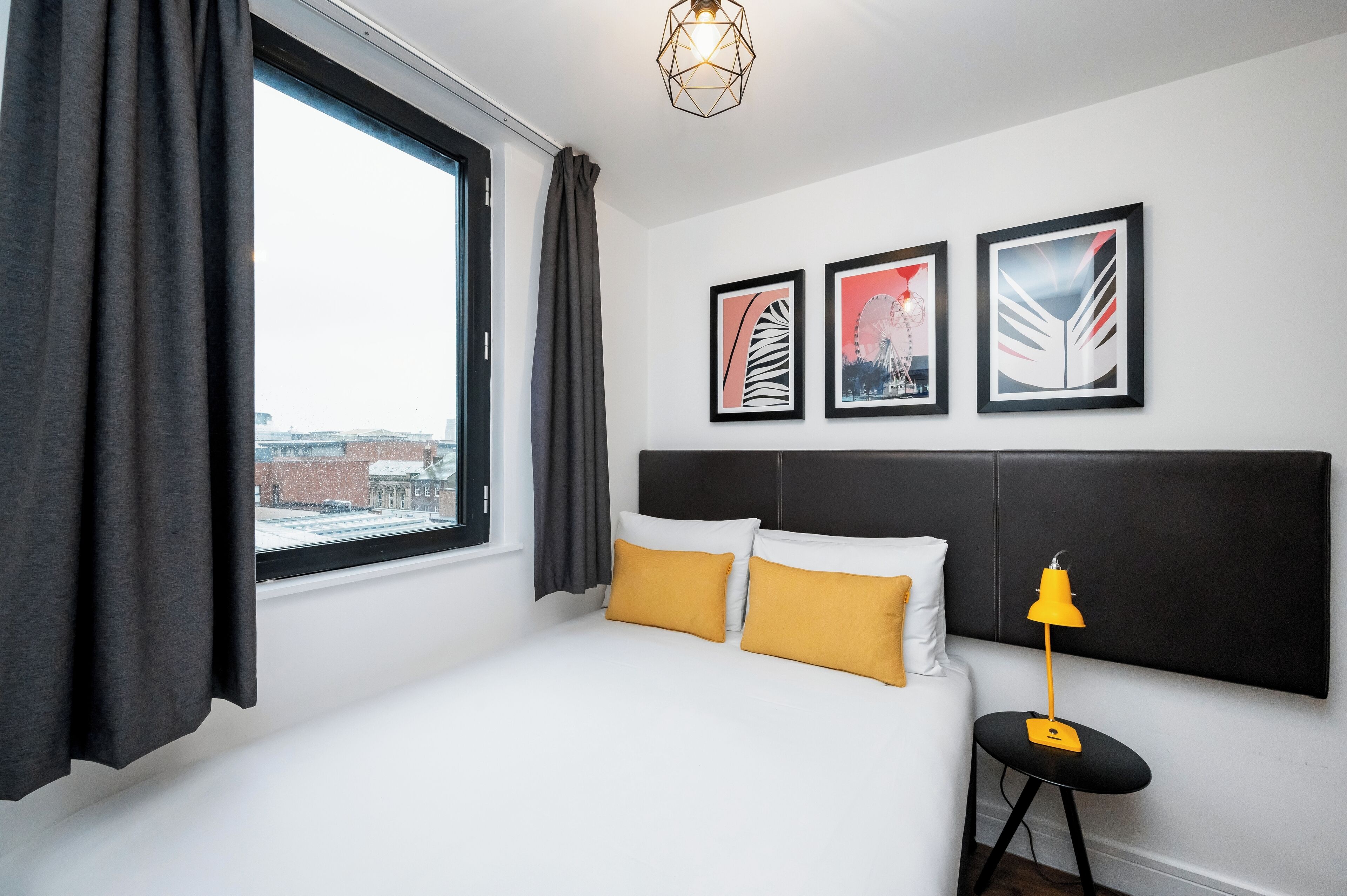 A hotel room with a large window, a bed with white and mustard pillows, a black headboard, and three framed artworks on the wall.