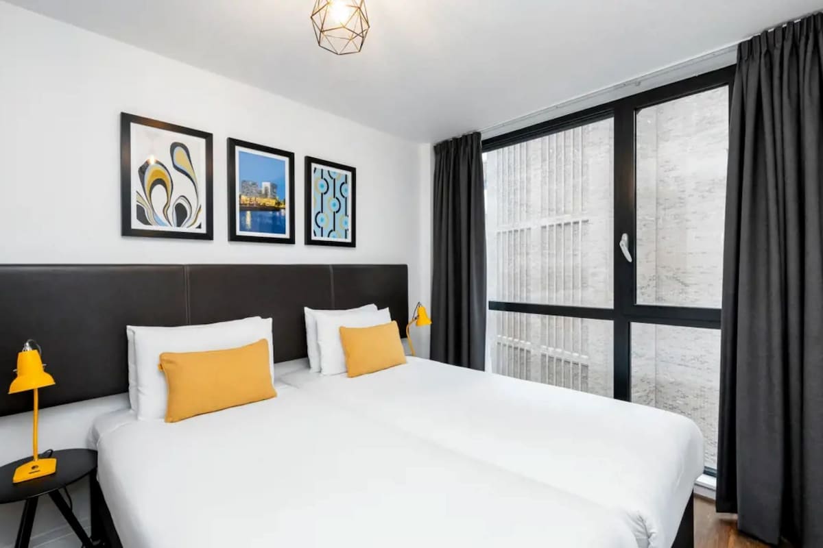 A modern hotel room with a large bed, two bedside tables with lamps, a large window with curtains, and three framed pictures on the wall.