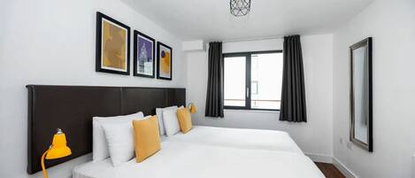 A modern hotel room with a large bed, a desk with a chair, a desk lamp, and a window with curtains.