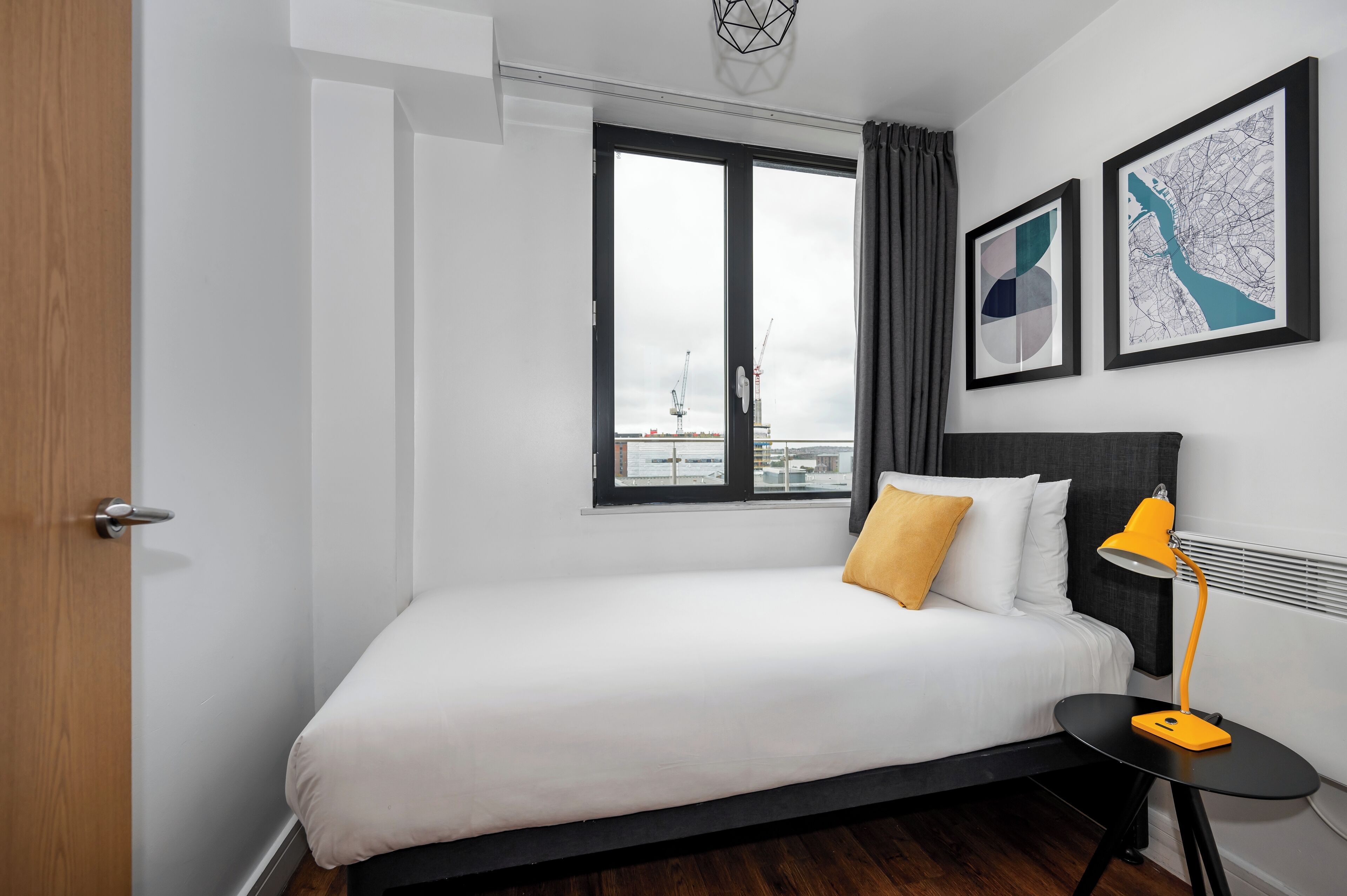 Ein modernes Hotelzimmer mit einem Bett, einem kleinen Tisch mit Lampe, einem Fenster mit Vorhängen und zwei gerahmten Kunstwerken an der Wand.