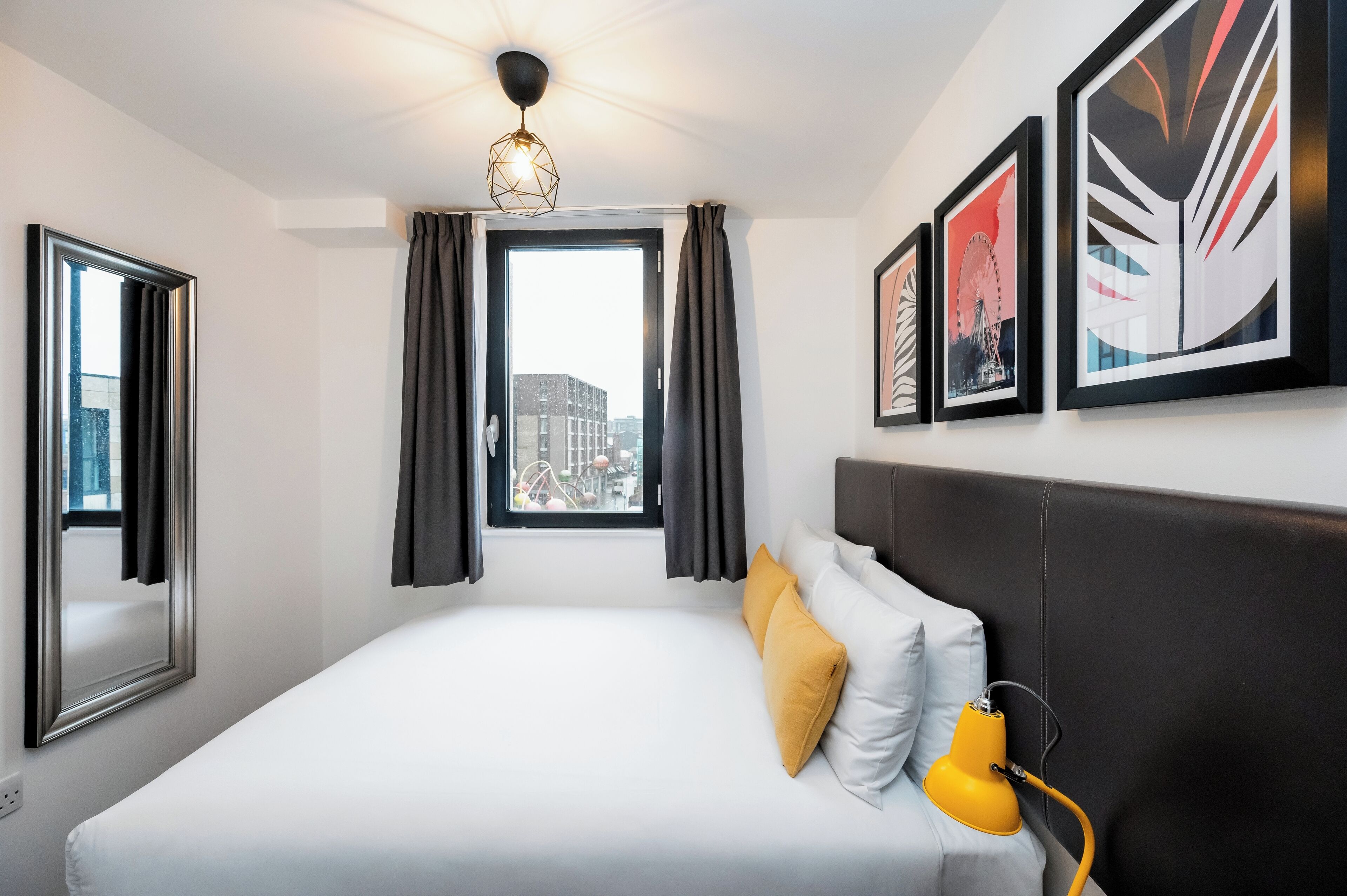 A hotel room with a bed, a window with curtains, a city view, and framed artwork on the wall.