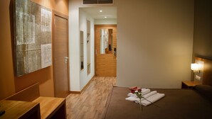 Una habitación de hotel con suelo de madera, una cama con cabecera marrón, una mesita de noche y una lámpara fijada a la pared.