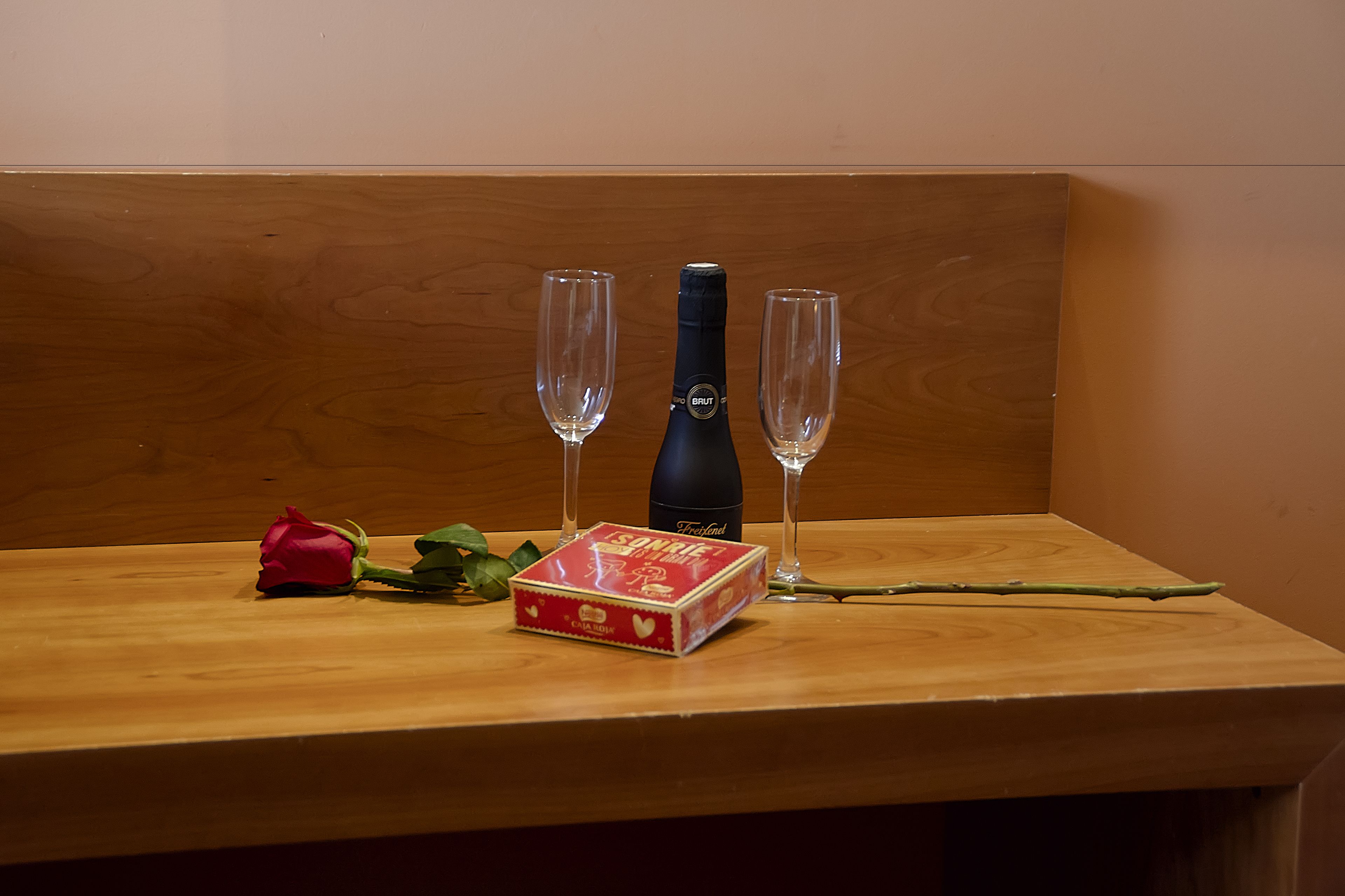 A wooden table with a bottle, two glasses, a red rose, and a gift box.