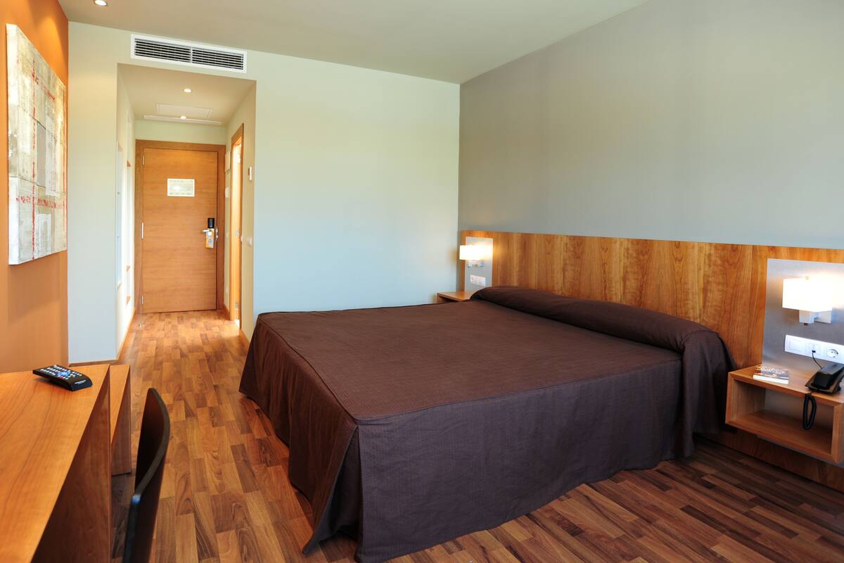A hotel room with a large bed, a desk with a chair, a bedside table with a lamp, and a wall-mounted phone.