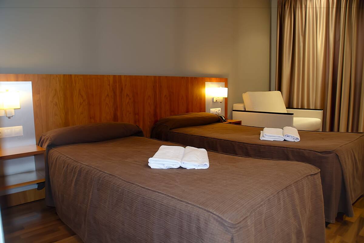 A hotel room with two beds, each with a brown bedspread and a folded towel on the bed. There is a nightstand with a lamp and a chair with a white cushion.