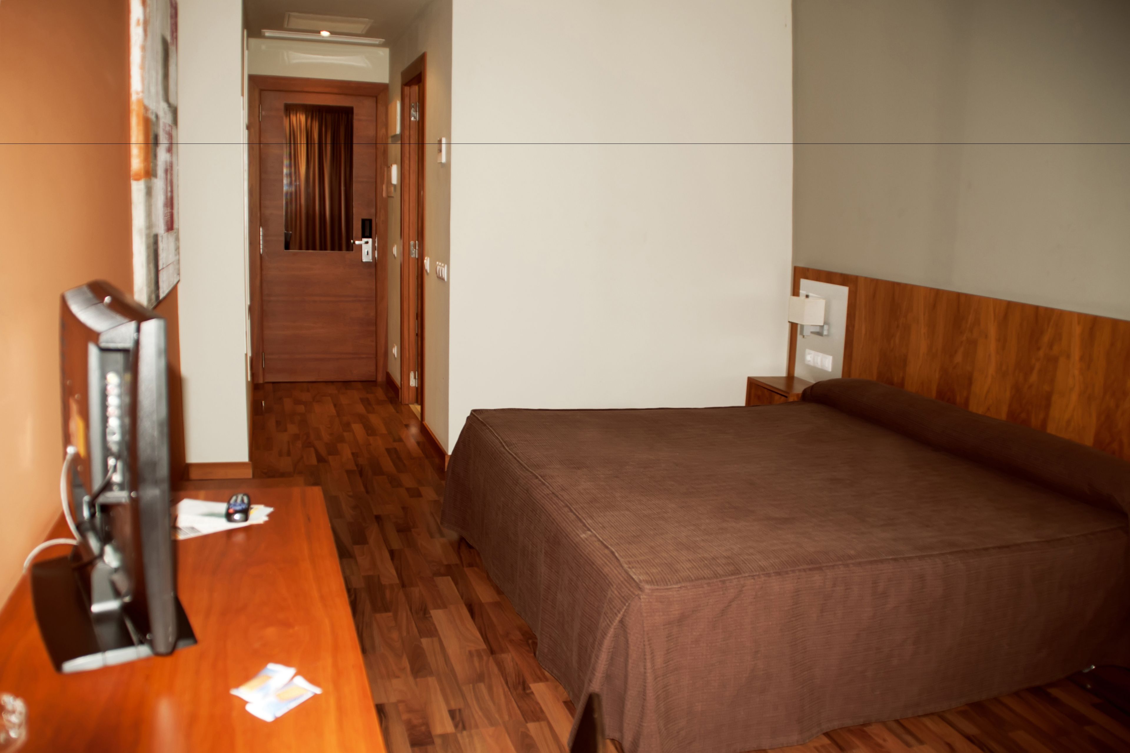 Habitación de hotel con suelo de madera, cama con cubrecama marrón, escritorio con computadora y puerta que da a otra habitación.
