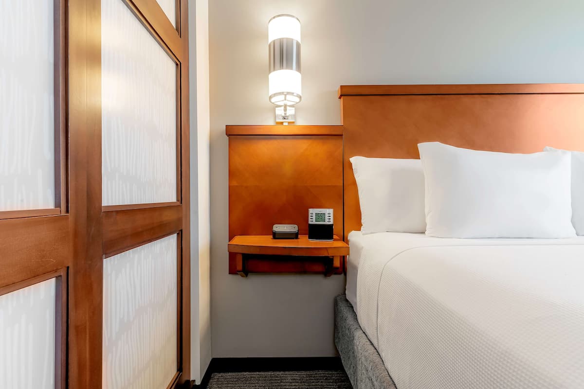 A hotel room with a wooden headboard, a bedside table with a clock, and a wall-mounted light fixture.