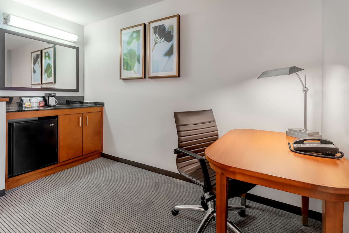 A modern hotel room with a wooden desk, a chair, a desk lamp, a telephone, a coffee maker, and a refrigerator.