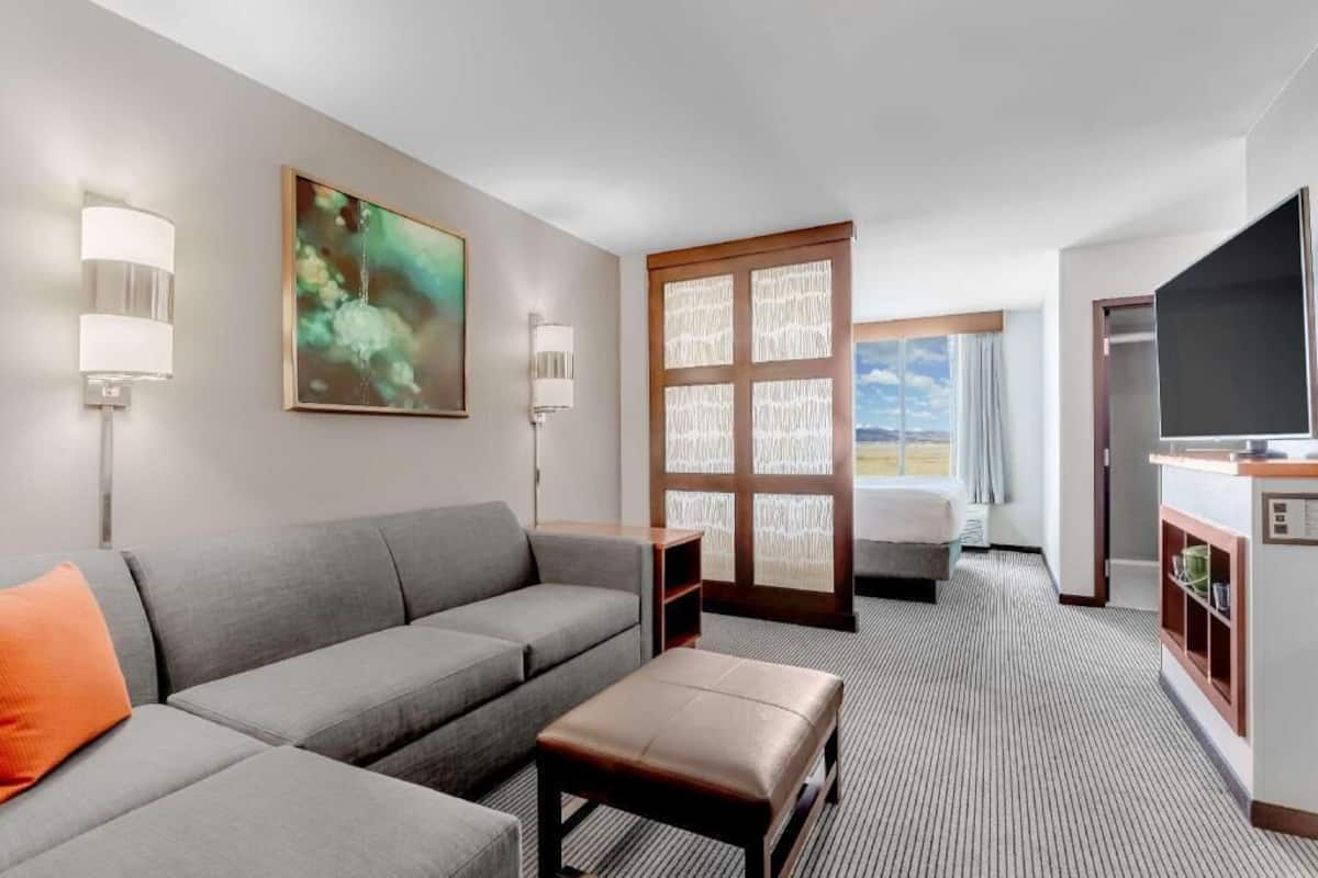 A hotel room with a grey sofa, a small table, a TV, a window with a view, and a door leading to another room.
