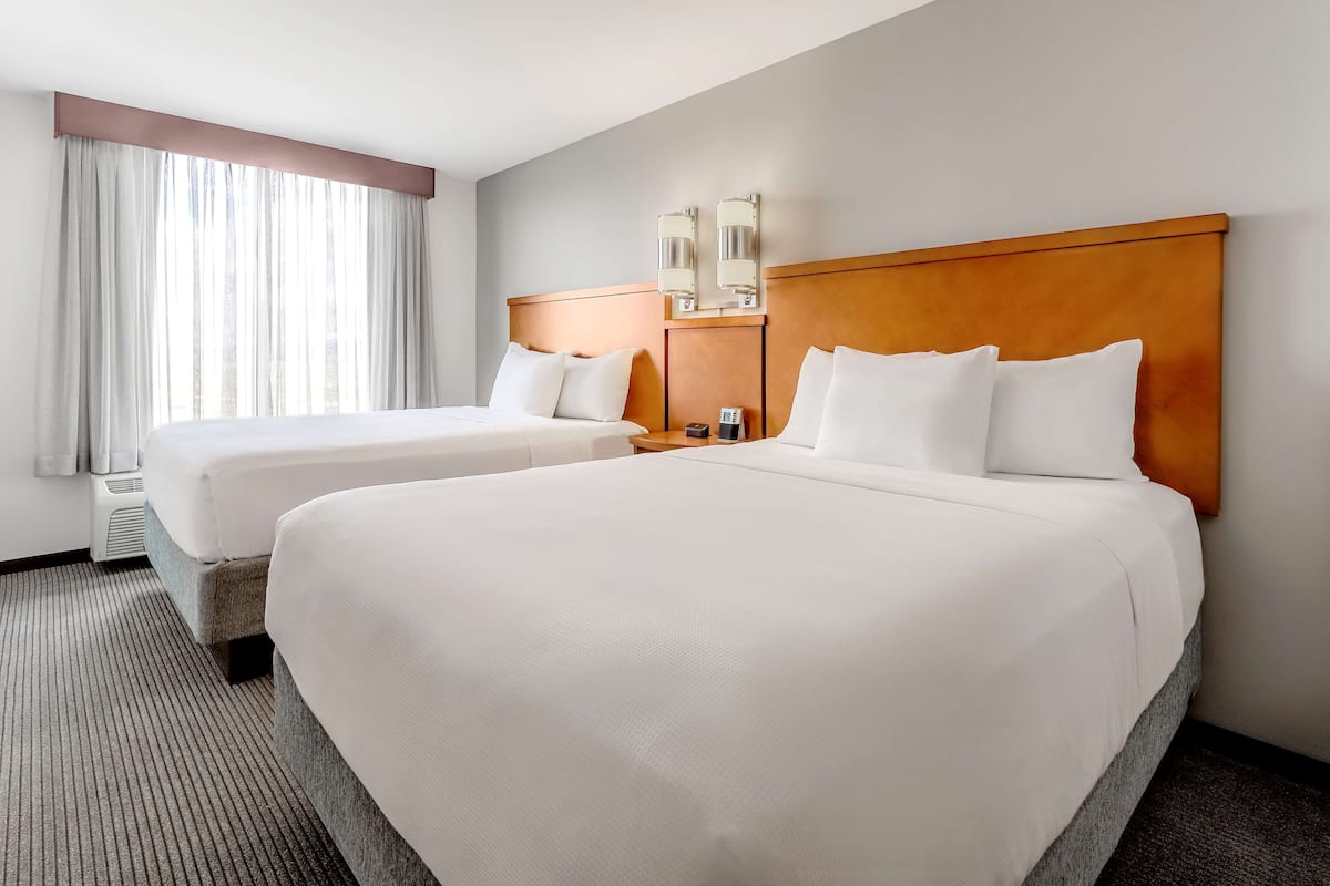 A hotel room with two beds, a large window with sheer curtains, a wooden headboard, and a wall-mounted light fixture.