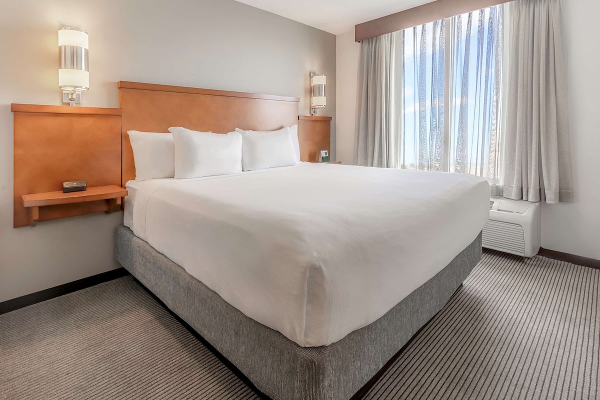A large bed with a wooden headboard, two bedside tables with lamps, a large window with curtains, and a carpeted floor.