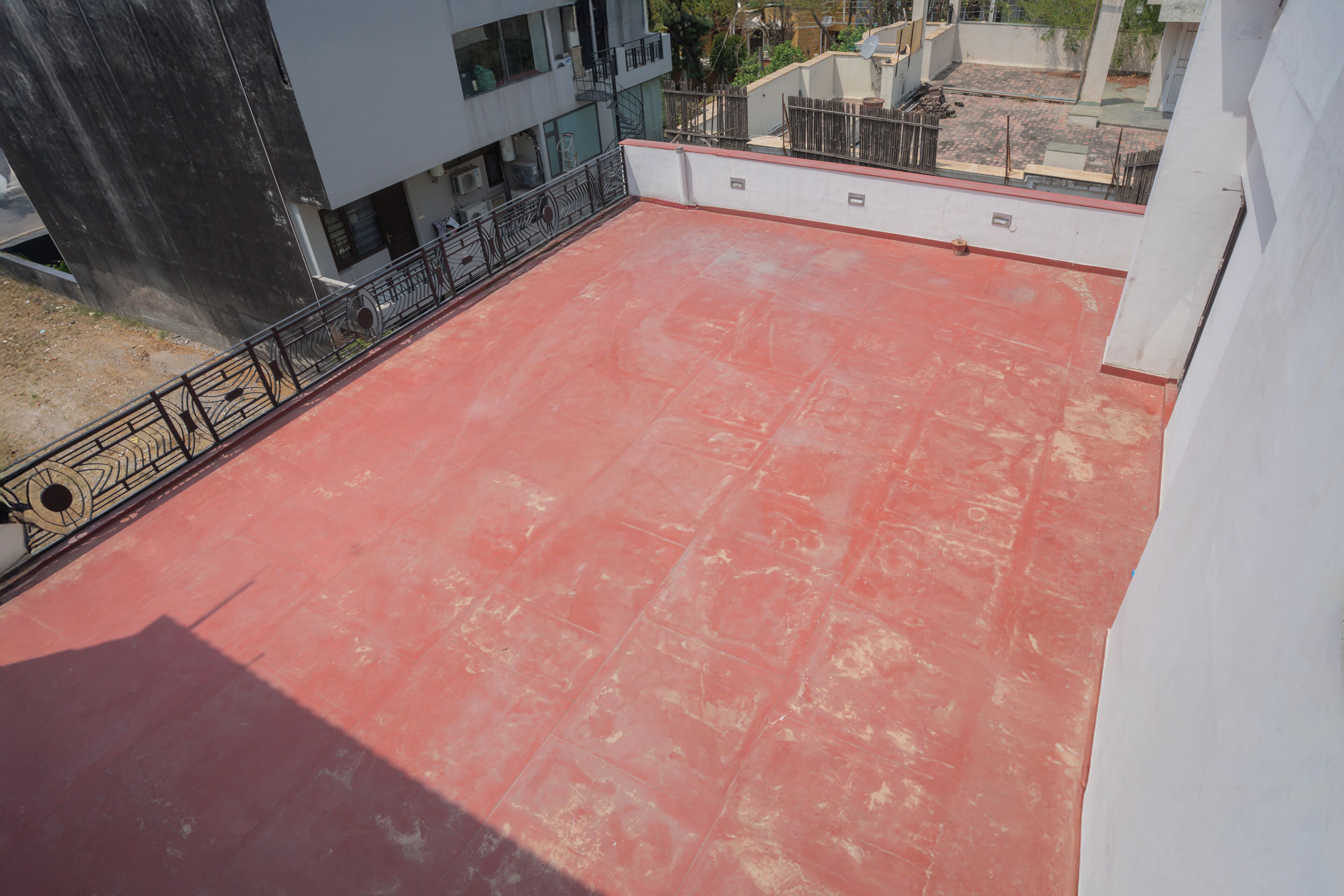 A rooftop terrace with a red surface, metal railing, and a view of surrounding buildings.