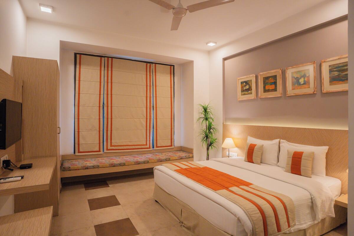 Una habitación de hotel moderna con una cama grande, un espacio de descanso con un banco, un televisor y obras de arte en las paredes.