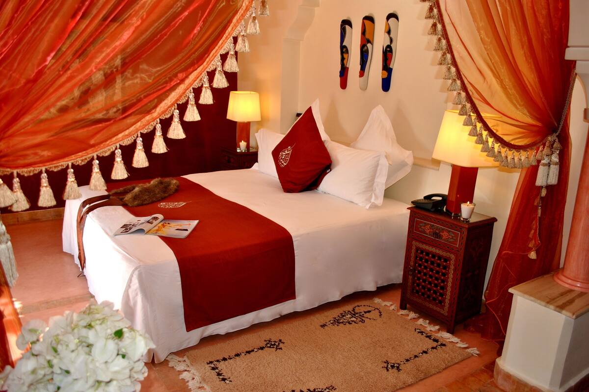 A neatly made bed with a red throw, white pillows, and a red accent pillow. There is a small wooden nightstand with a lamp and a decorative item.
