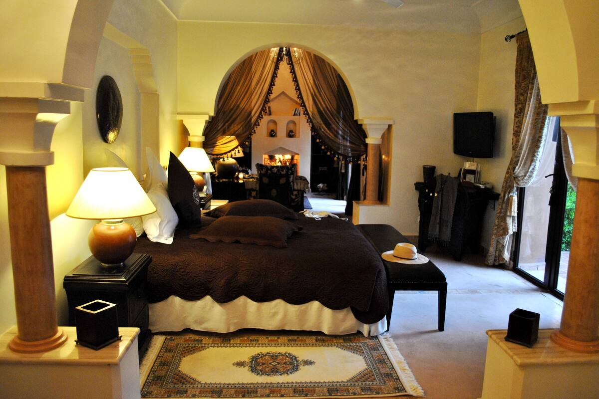 A bedroom with a large bed, a nightstand with a lamp, a small table with a candle, and a view of a courtyard through a window.