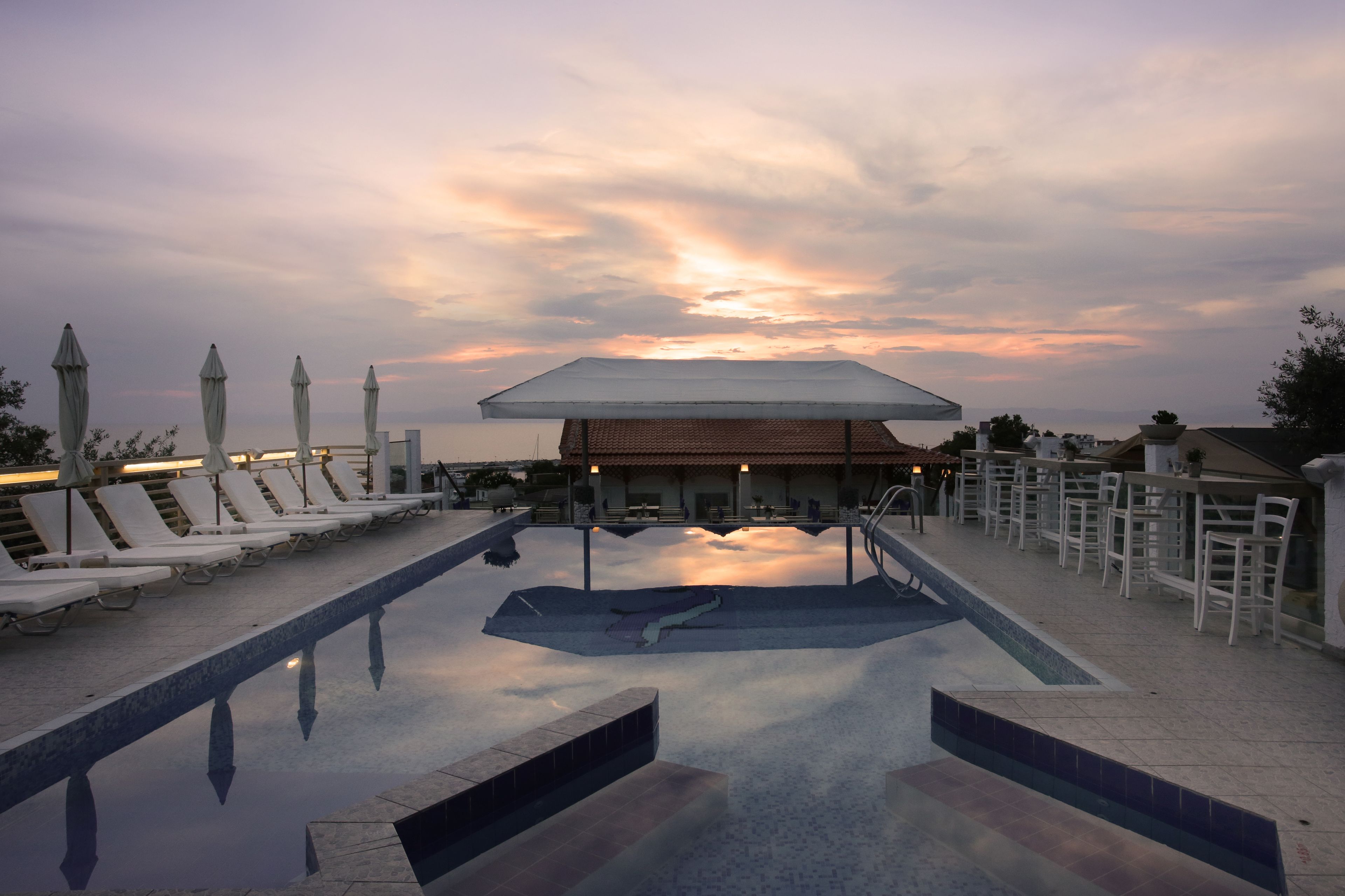 Zona de piscina con sillas de descanso y sombrillas, un espacio de estar cubierto y vistas a la puesta de sol.