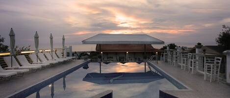 Zona de piscina con sillas de descanso y sombrillas, un espacio de estar cubierto y vistas a la puesta de sol.