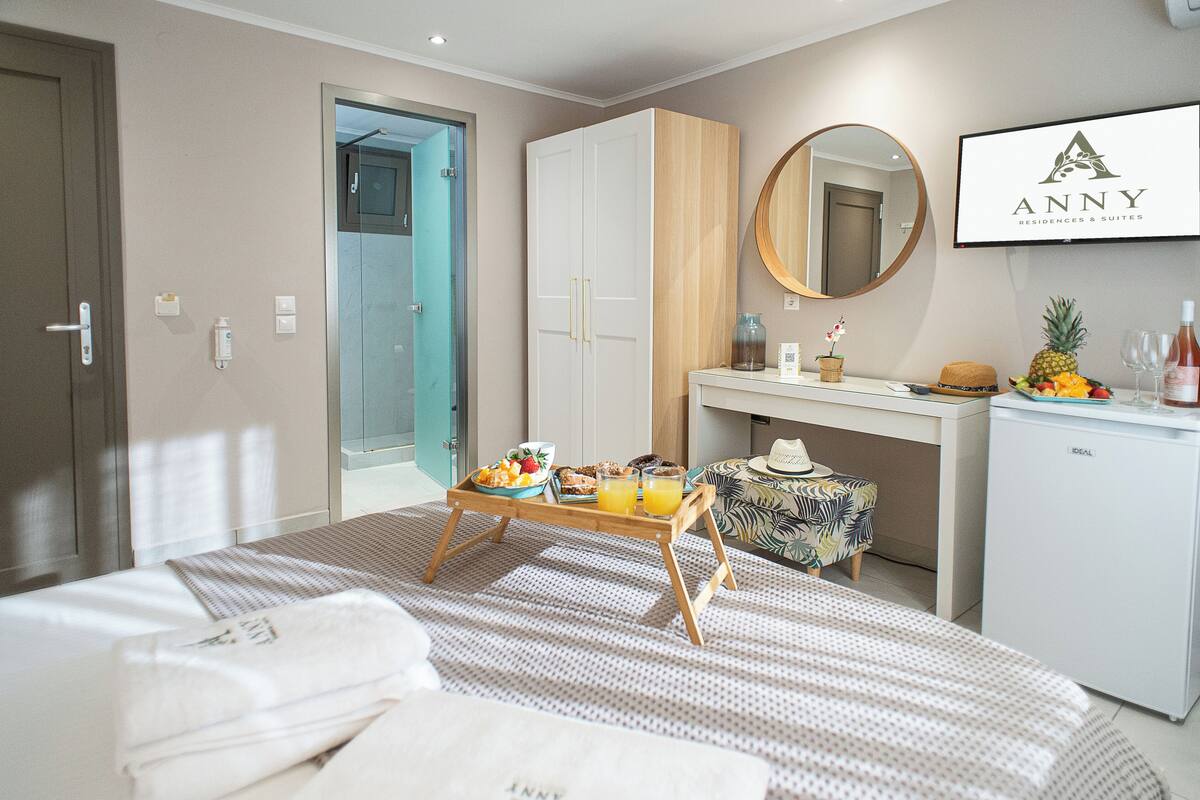 A hotel room with a bed, a wooden wardrobe, a desk with a mirror, a small fridge, and a breakfast tray on the bed.