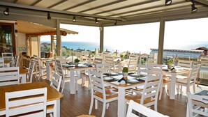 A restaurant with white chairs and tables, large windows, and a view of the sea. - Anny Residences & Suites (Thasos)