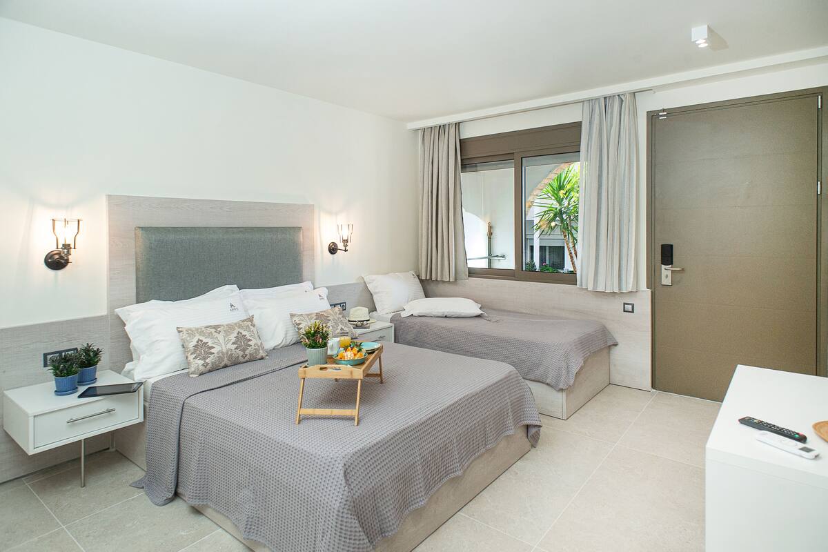 A modern hotel room with two beds, a bedside table with a lamp, a small desk with a chair, and a window with curtains.