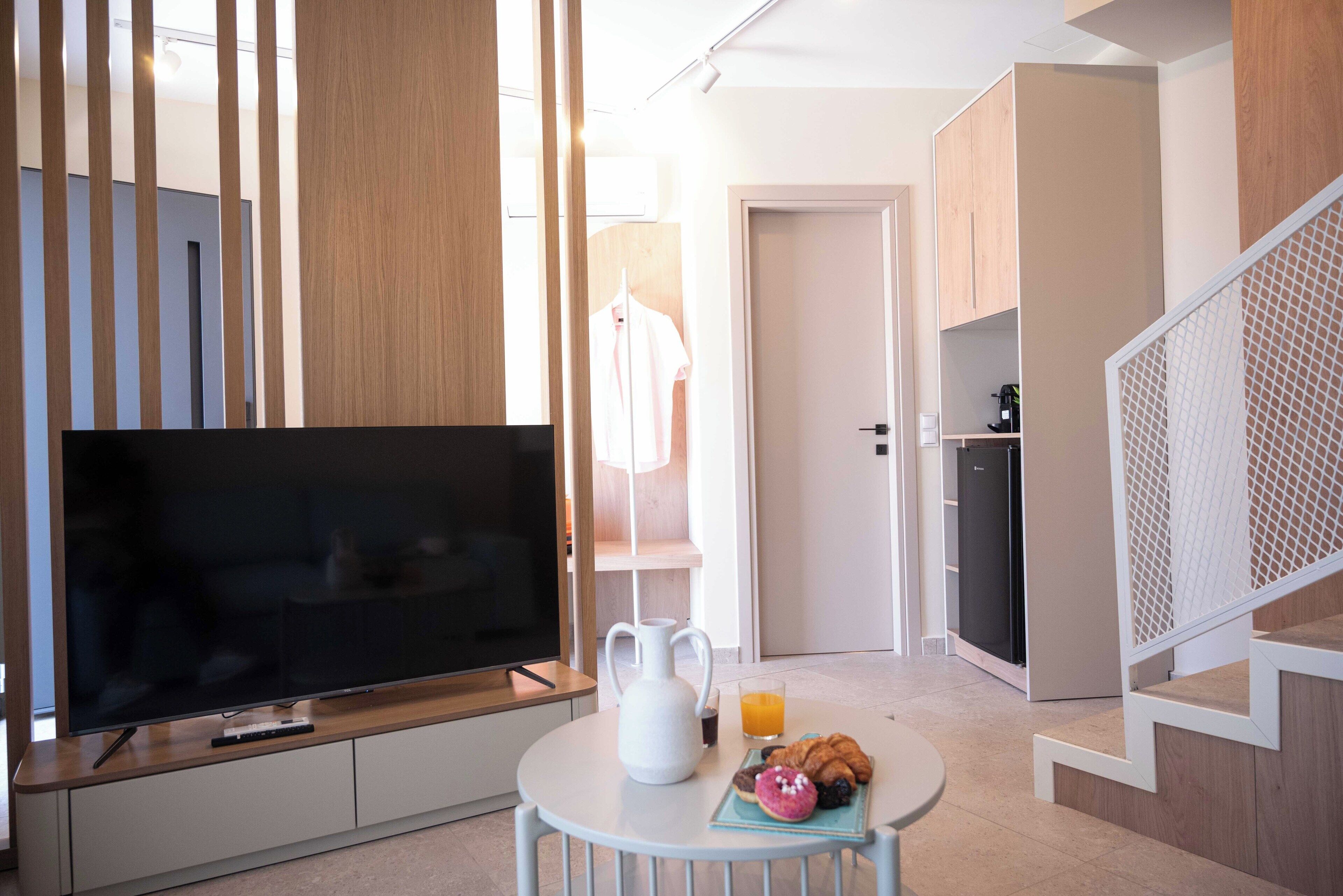 Una habitación de hotel moderna con un televisor de pantalla plana, una jarra blanca, un vaso de jugo de naranja y un plato de croissants sobre una mesita.