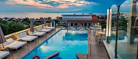 A rooftop pool with lounge chairs and umbrellas, a lifebuoy, and a view of the sea and buildings in the background.