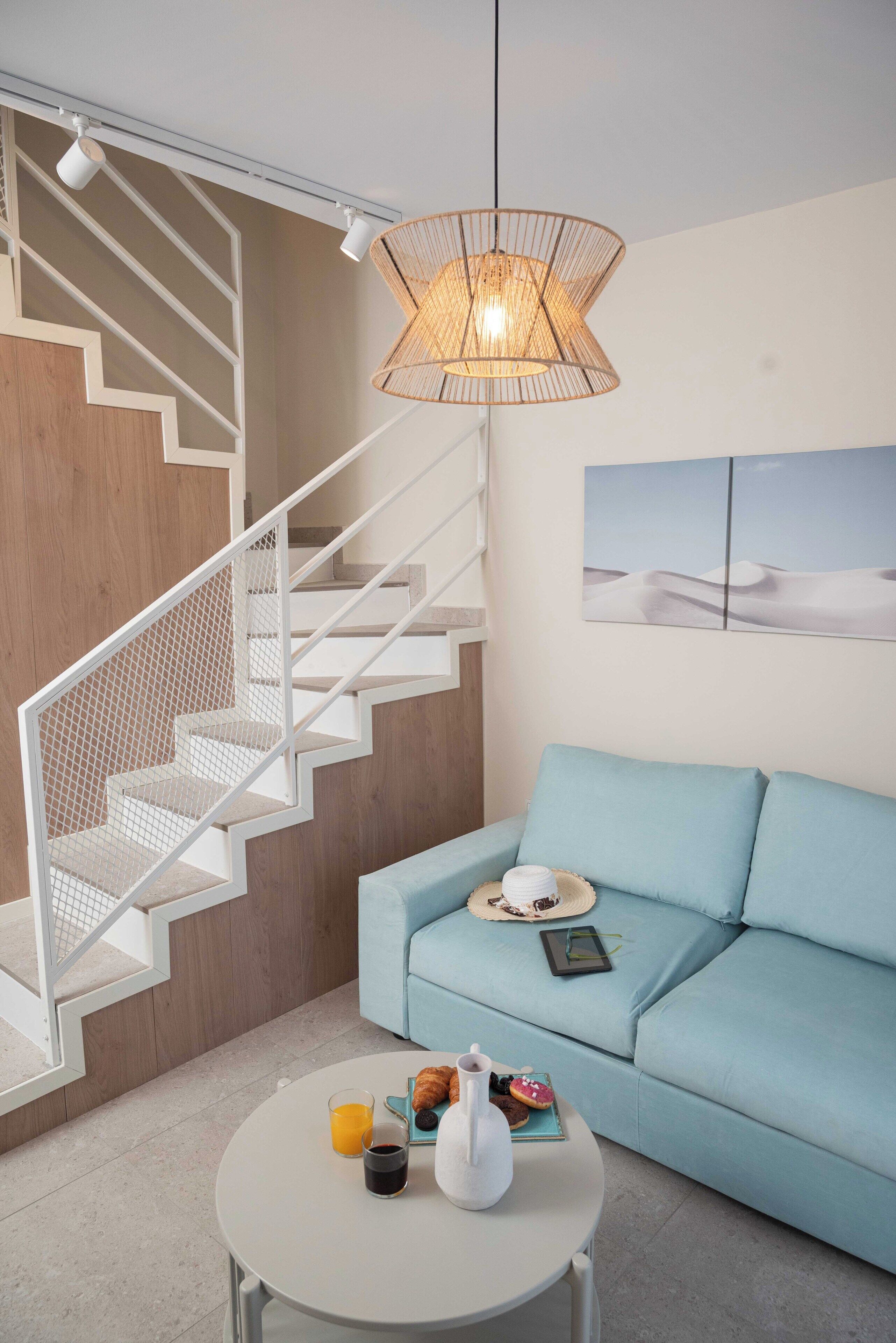 Una sala de estar moderna con una escalera, un sofá azul claro, una mesa redonda de centro con alimentos para el desayuno y una lámpara colgante.