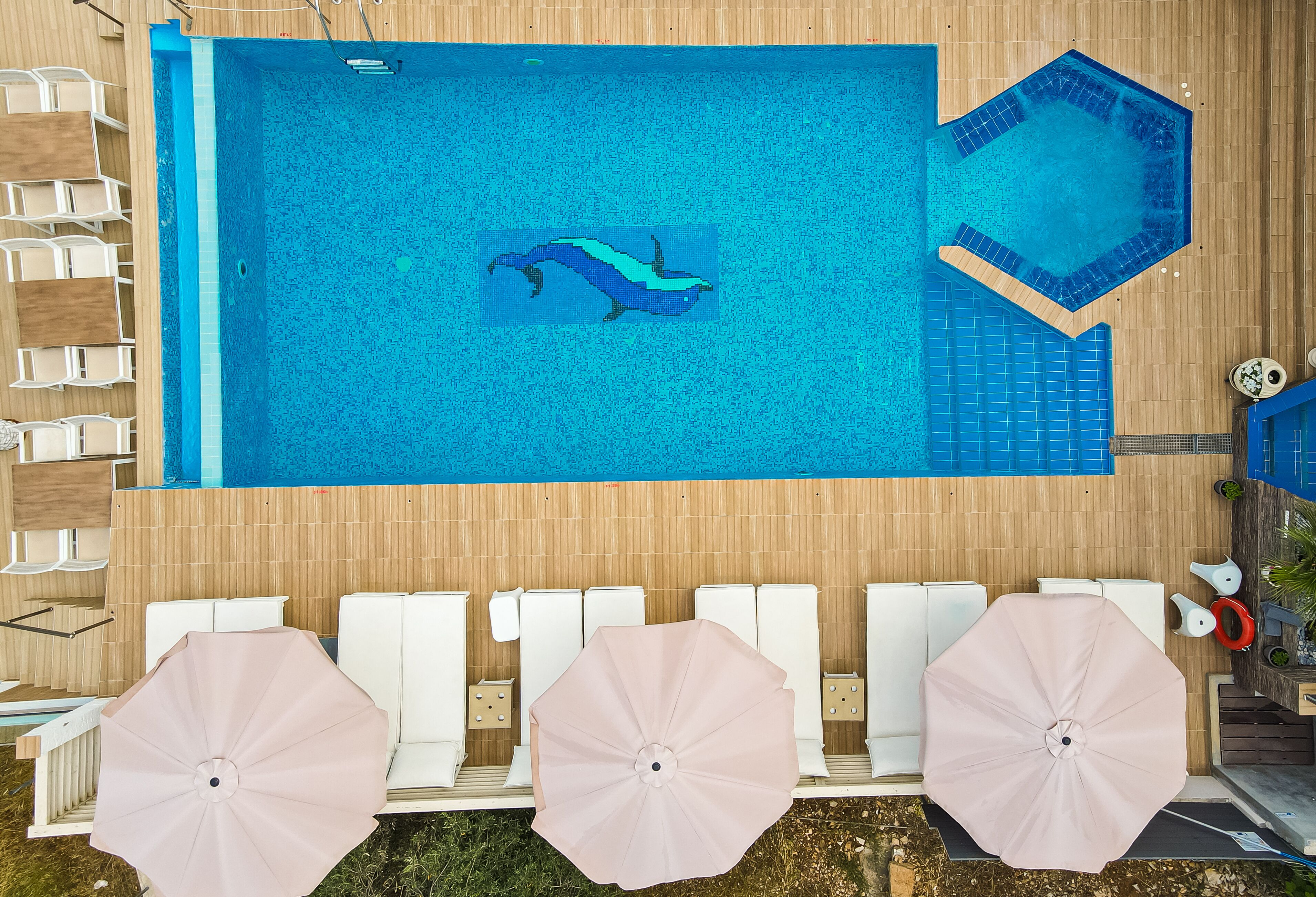 Una piscina con un mural de delfines, sillas de descanso blancas y sombrillas rosas.