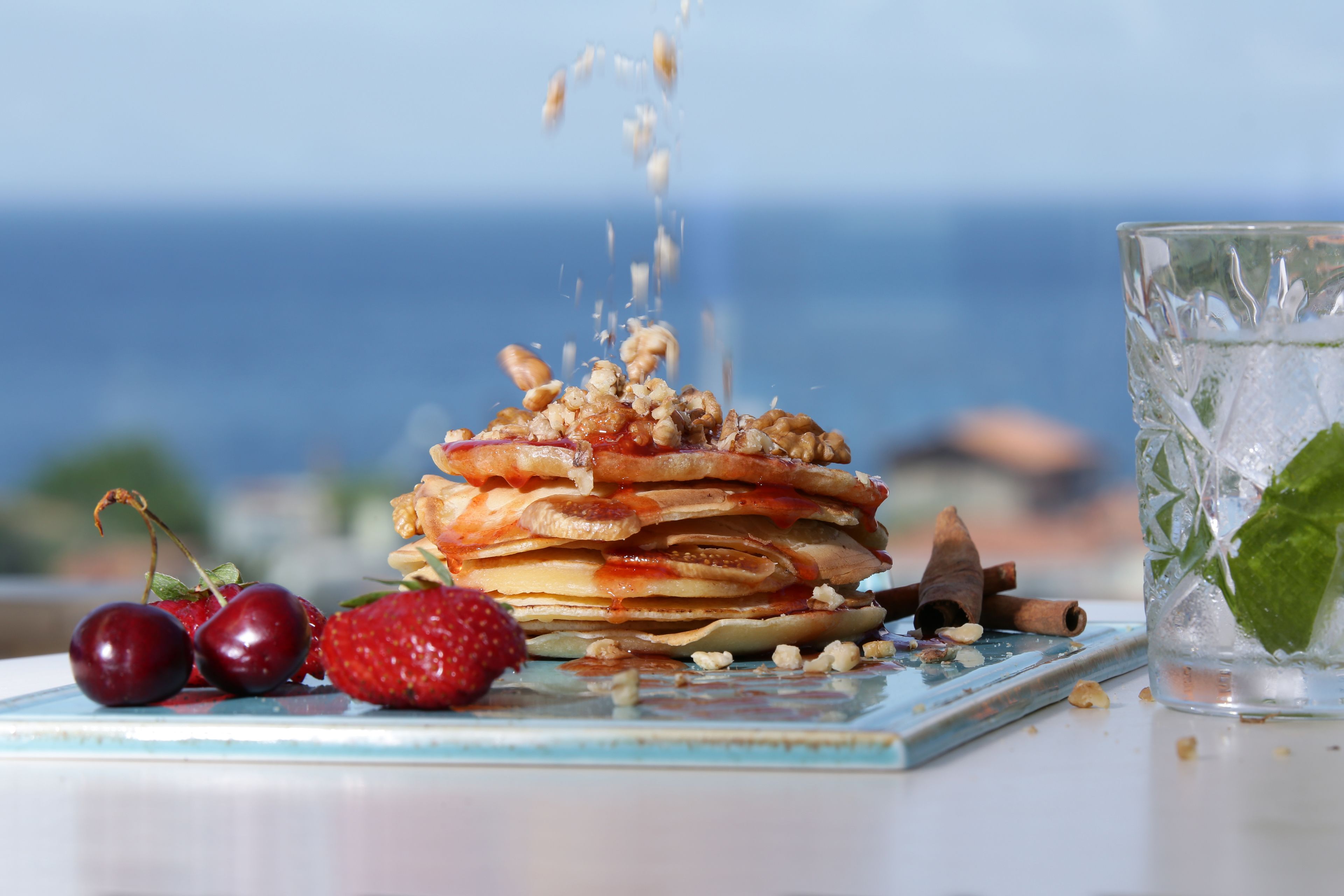 Une pile de crêpes garnie de cerises et de noix, présentée sur une assiette, à côté d’un verre d’eau glacée avec des feuilles de menthe.