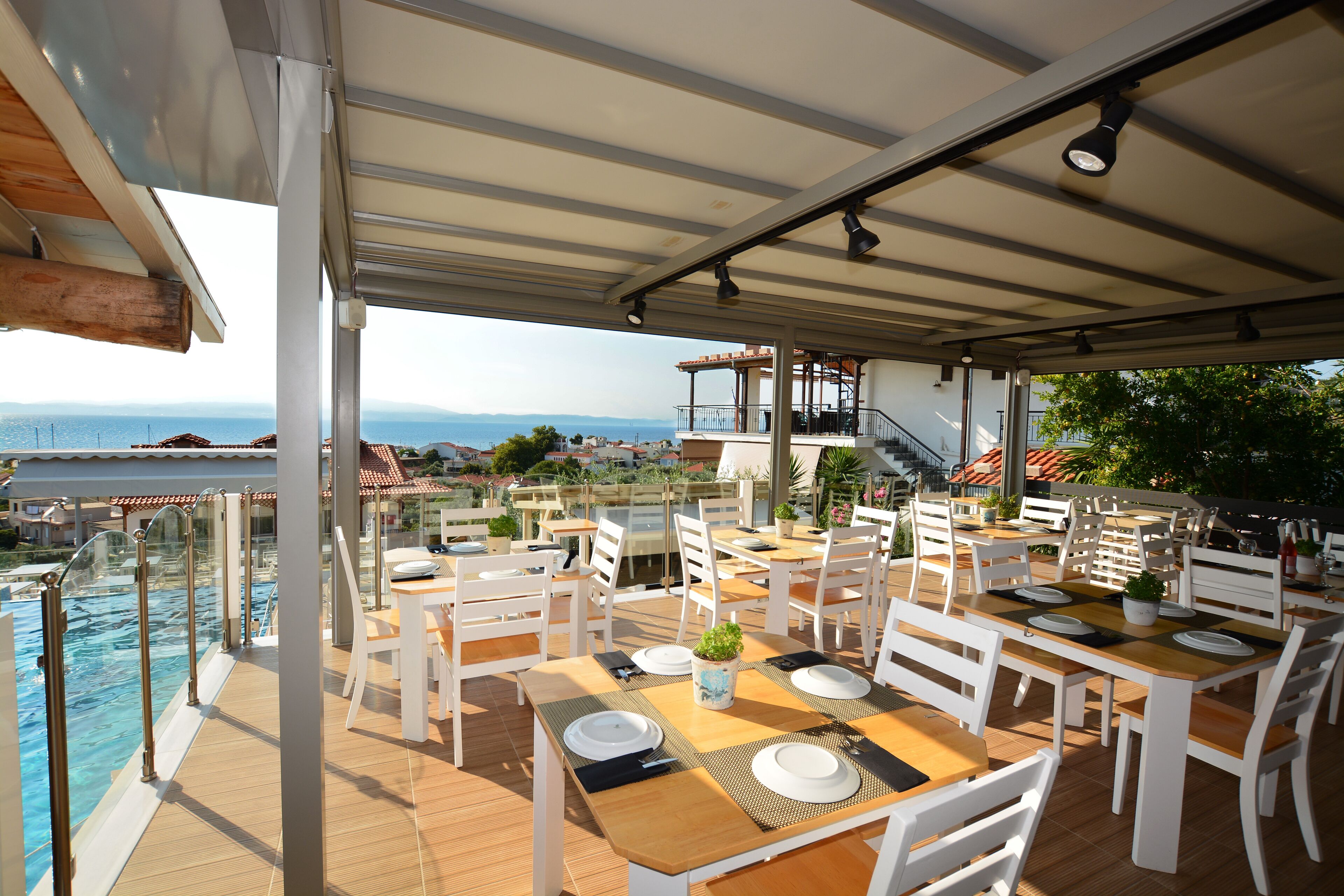 An outdoor dining area with tables and chairs, overlooking a pool and a scenic view of the sea and mountains.