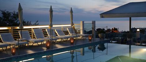 Ein Poolbereich mit Liegestühlen und Sonnenschirmen, ein klares Schwimmbecken und Blick auf das Meer im Abendrot.