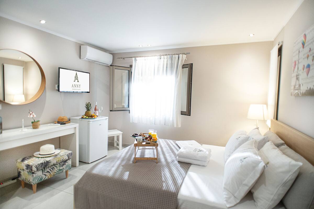 A hotel room with a bed, a desk, a chair, a small table with breakfast, a mirror, and a wall-mounted TV displaying the logo "ANNY".