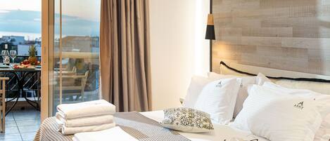 Habitación de hotel con cama, una mesa de comedor con sillas, vista a la ciudad y un balcón con una mesa dispuesta con frutas y vasos.