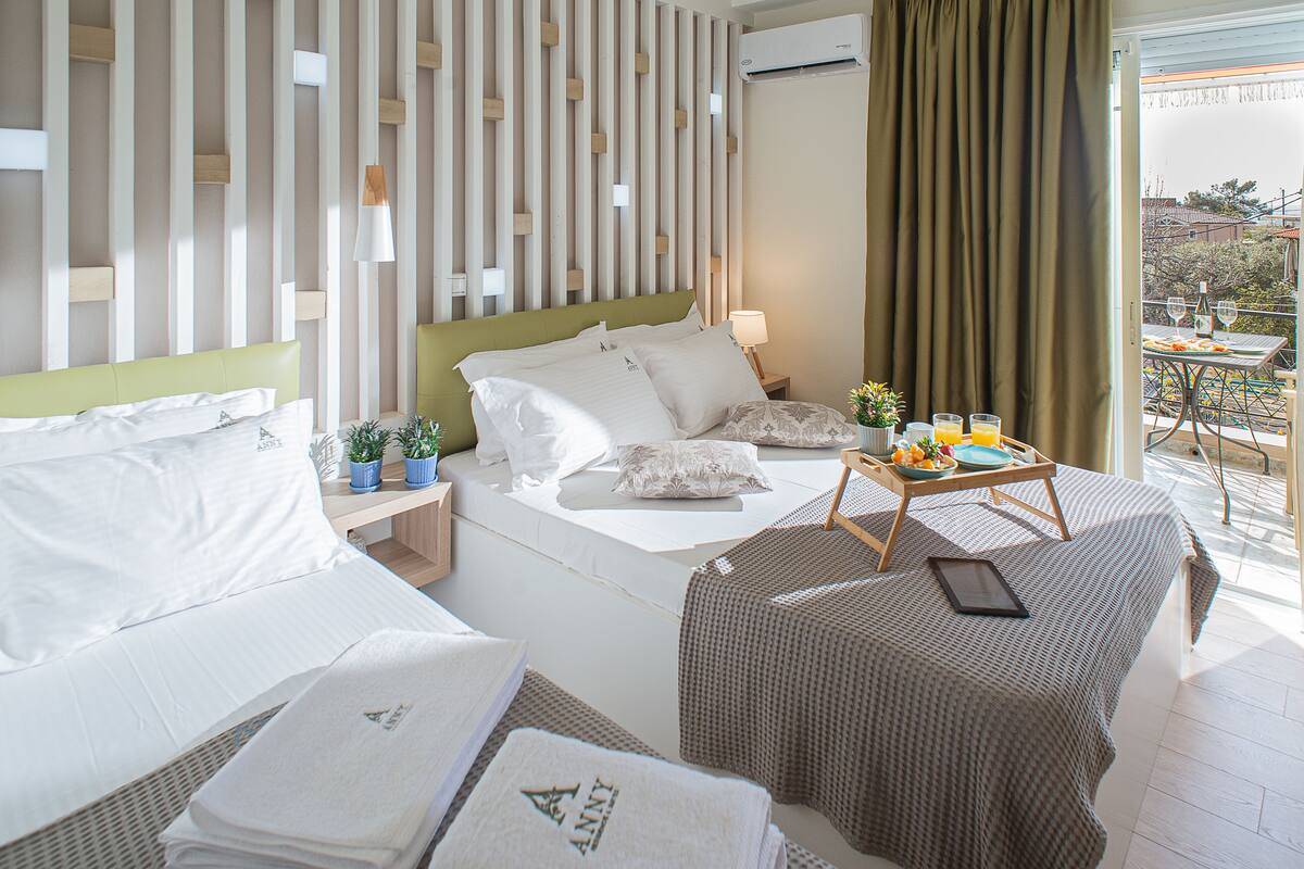 A hotel room with a bed, bedside table, a small table with a tray of fruit and glasses, a potted plant, and a view of the outside balcony.