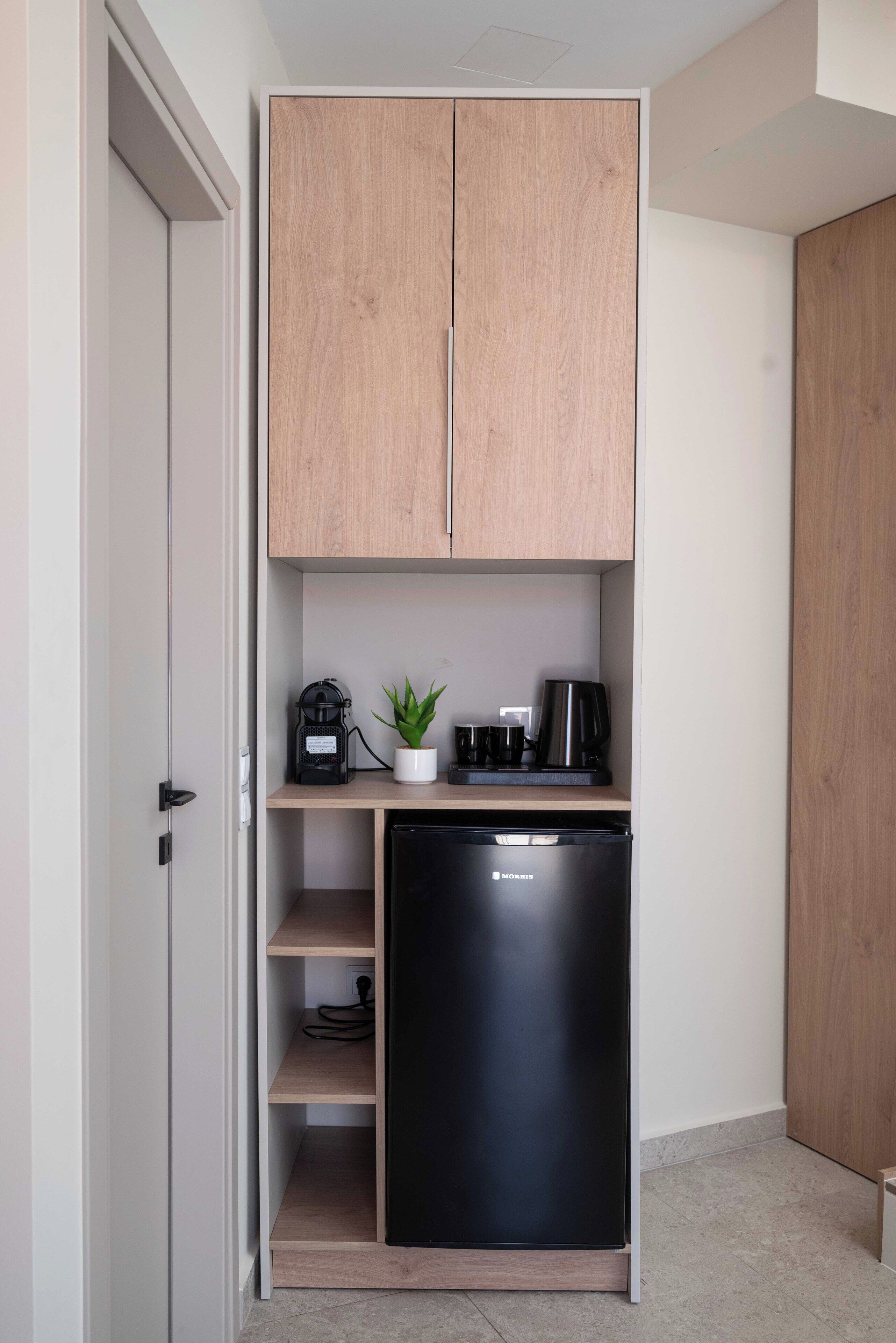 Una kitchenette compacta con un refrigerador negro, armarios de madera, un pequeño estante con una planta y una cafetera.