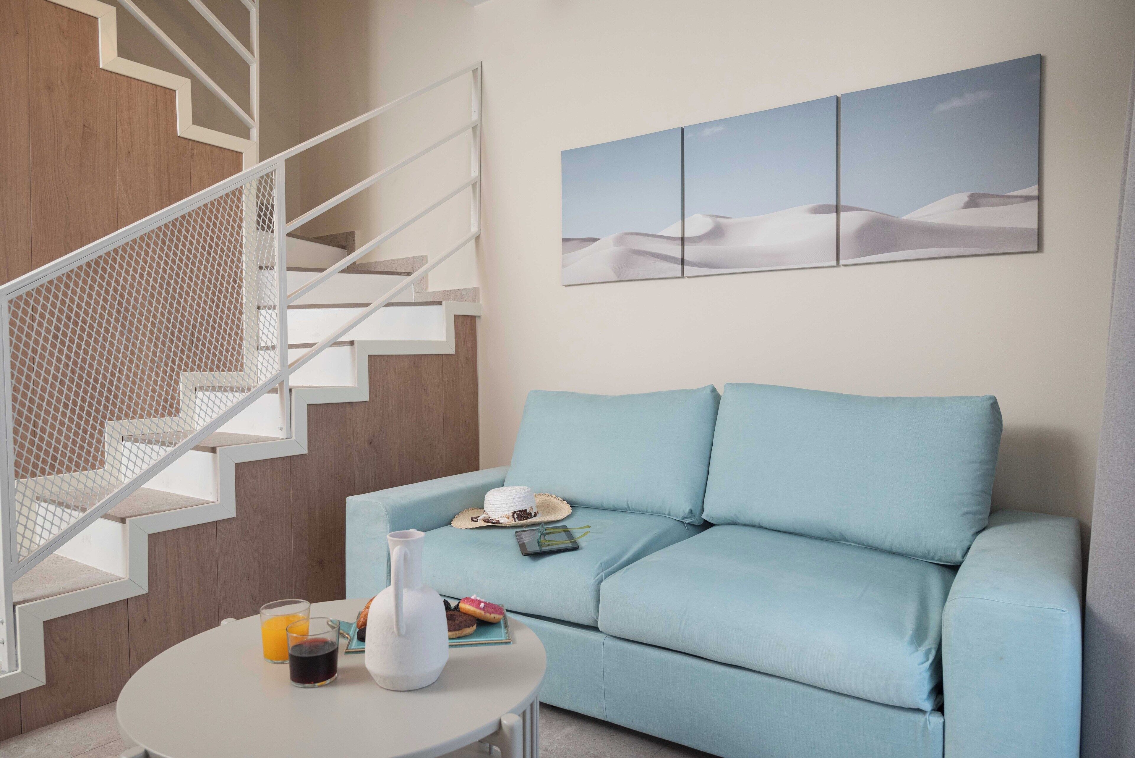 A modern living room with a light blue sofa, a white coffee table with drinks and a vase, a staircase with a white railing, and a desert landscape painting on the wall.