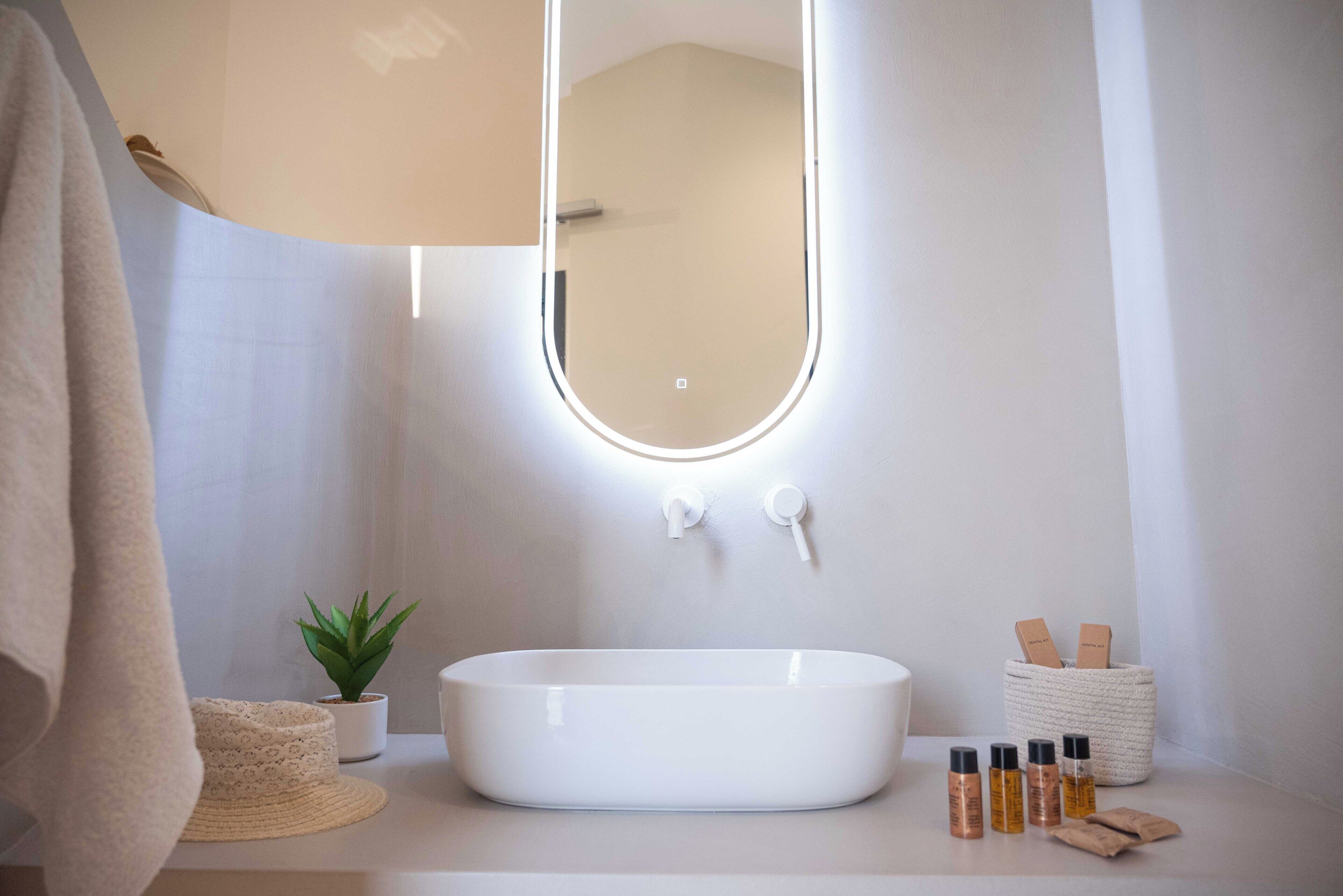 A modern bathroom with a white sink, a round mirror, a small potted plant, and a basket of toiletries.