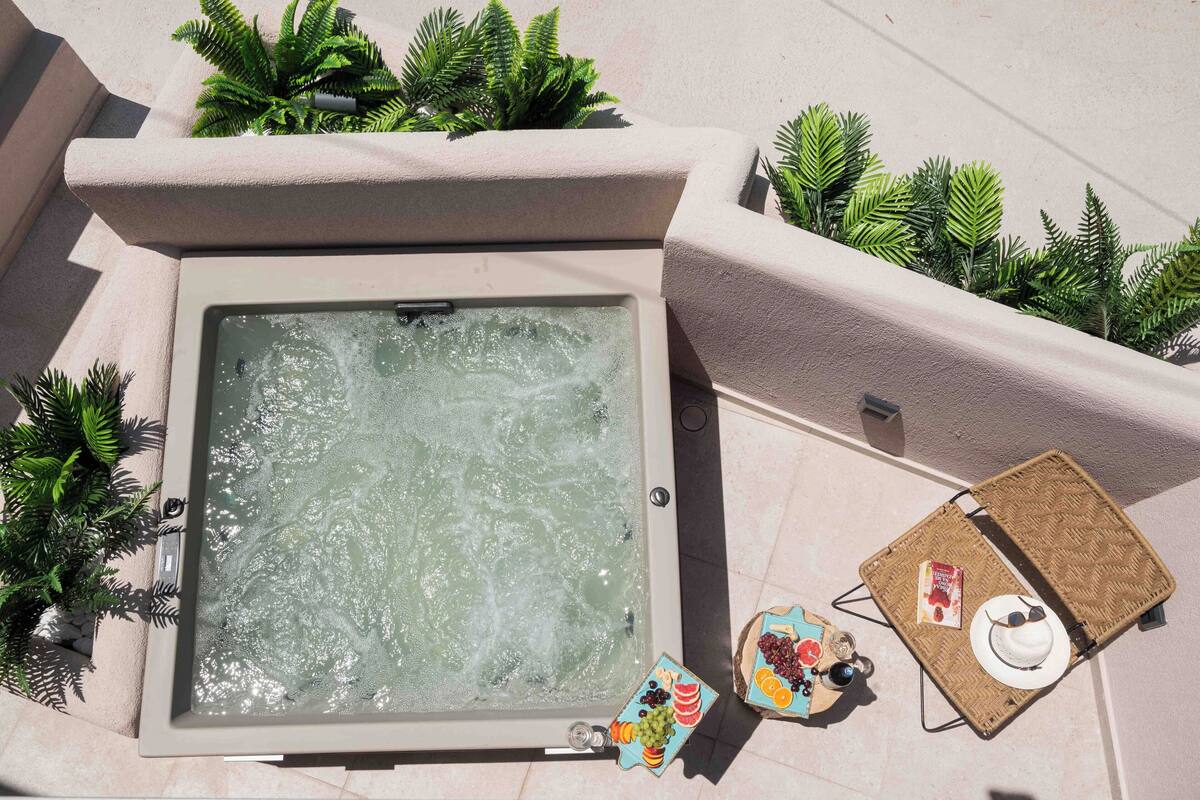 A hot tub with a view of the sky, surrounded by plants and a wicker chair with a hat and sunglasses.