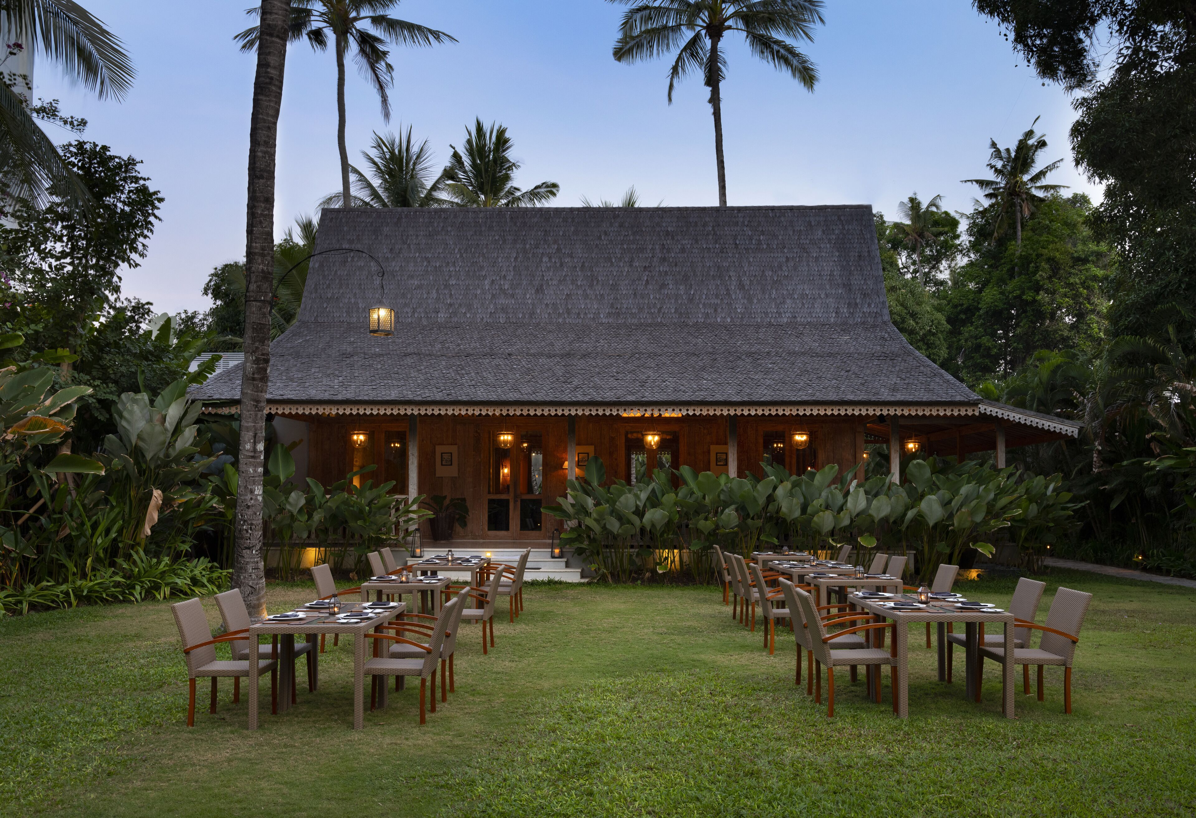 A restaurant with a thatched roof, outdoor seating, and surrounded by tropical vegetation.