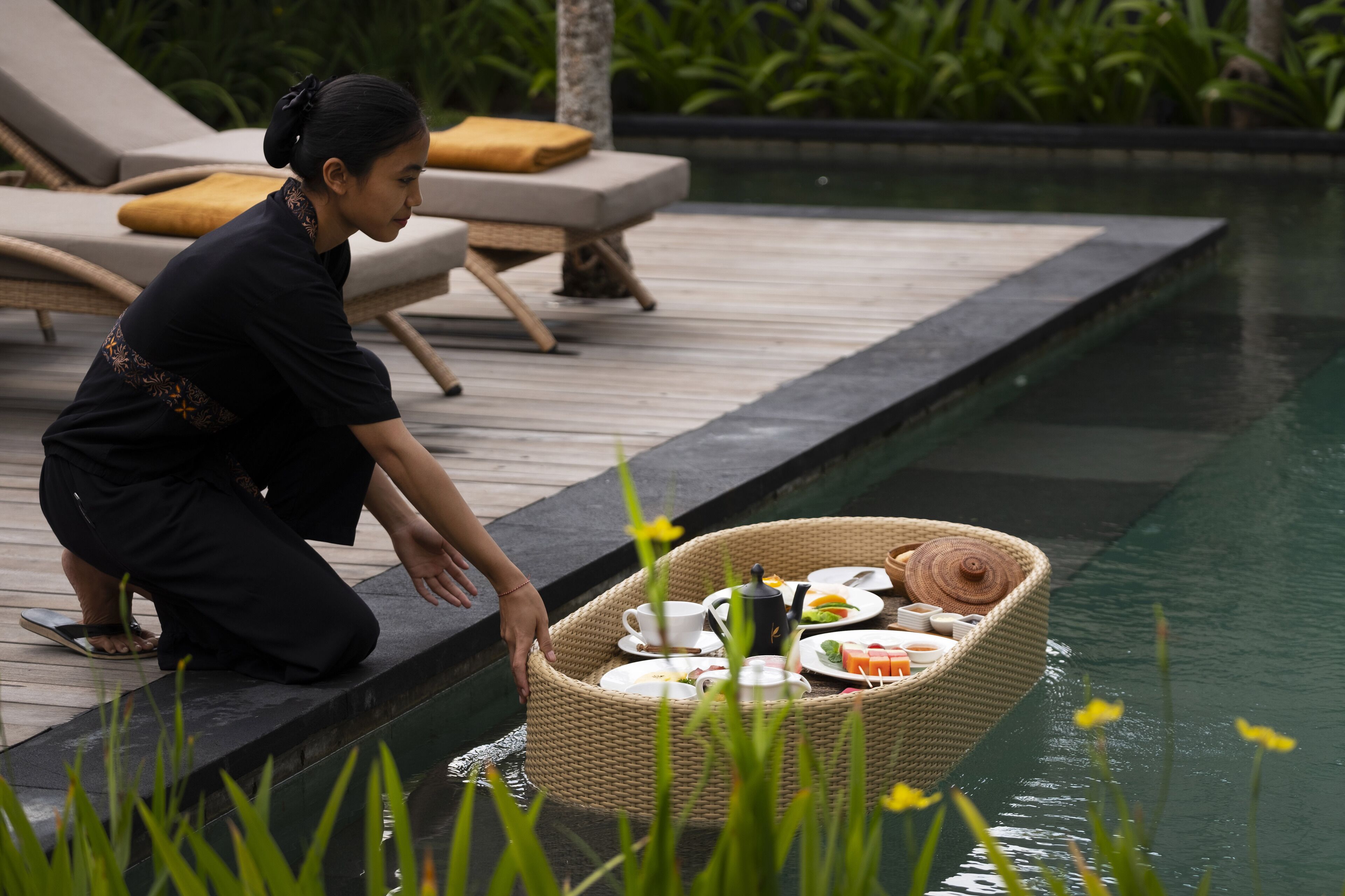 A person on a wooden deck by a pool, a wicker basket with food and drinks, and lounge chairs with cushions.