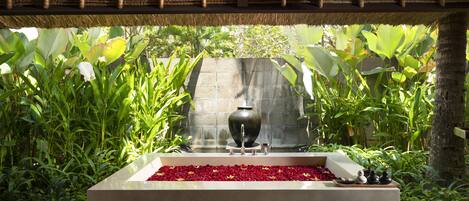 A rectangular stone bathtub with a black vase and a small fountain, surrounded by lush green plants and a stone wall.