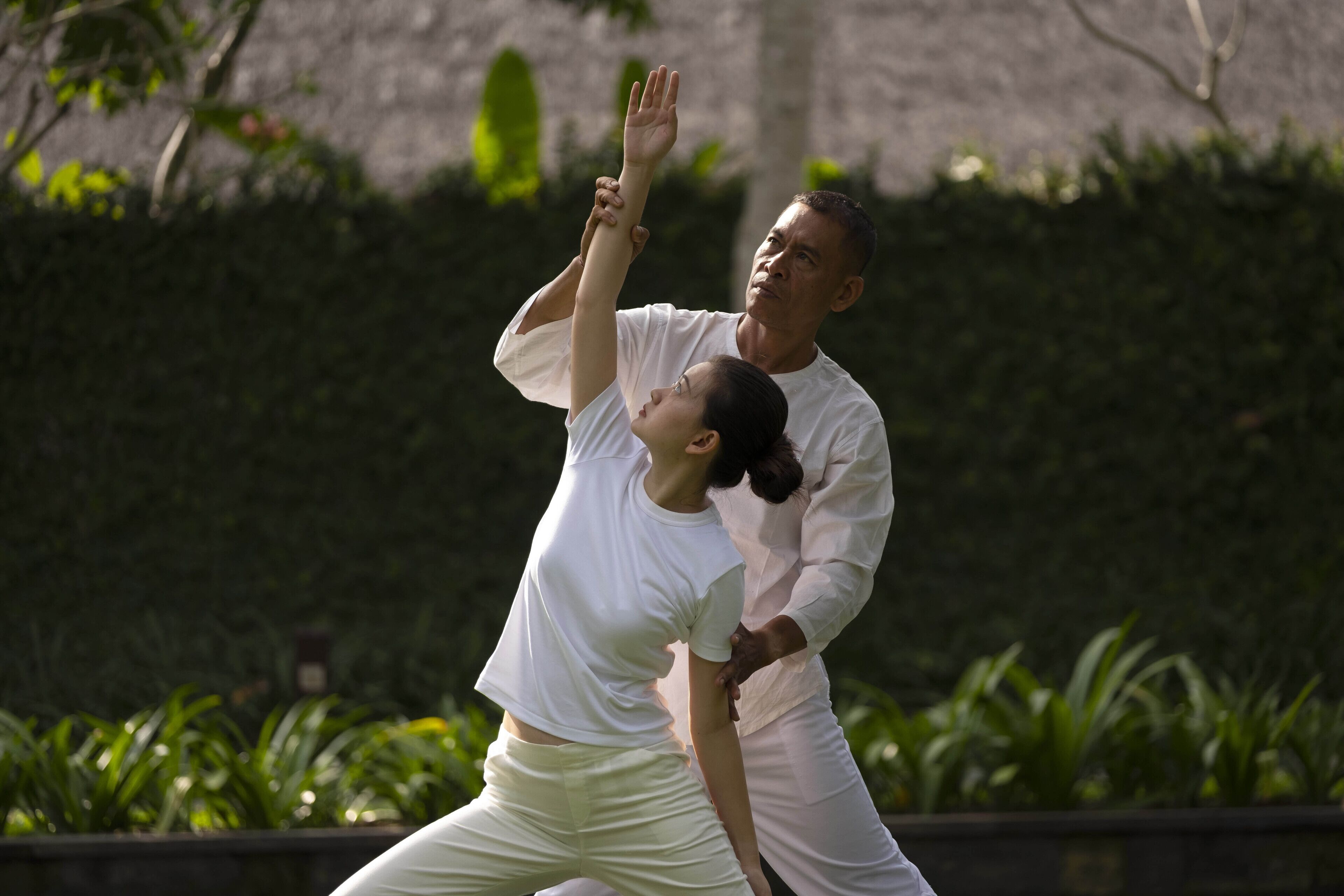 Una persona vestida de blanco realiza una técnica de artes marciales al aire libre, con vegetación de fondo.