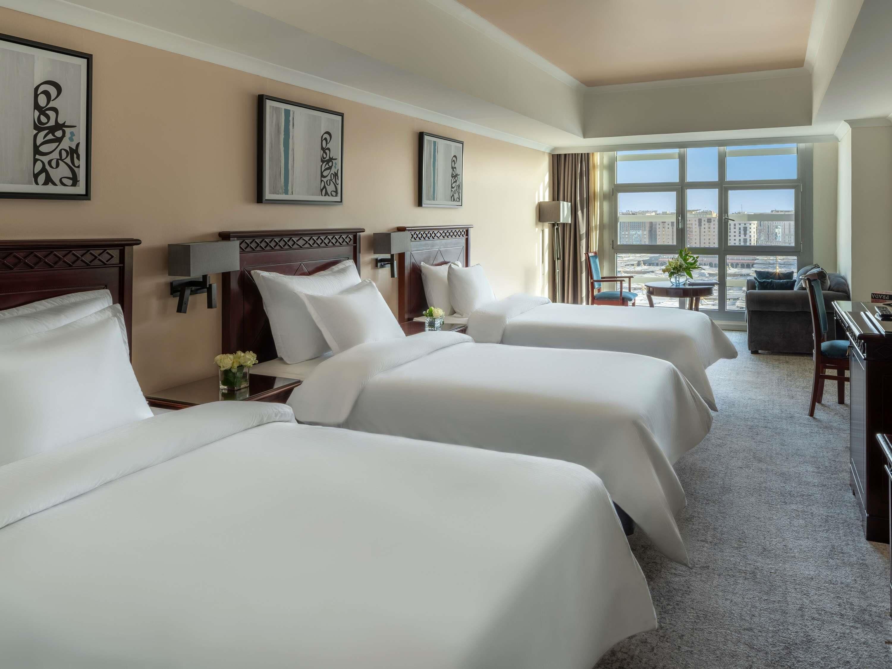 A hotel room with three beds, a desk, a chair, and a view of the city.