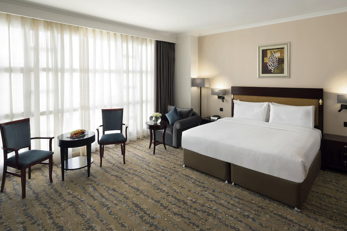 A hotel room with a large bed, two armchairs, a small table with a fruit bowl, a nightstand with a lamp, and a painting on the wall.
