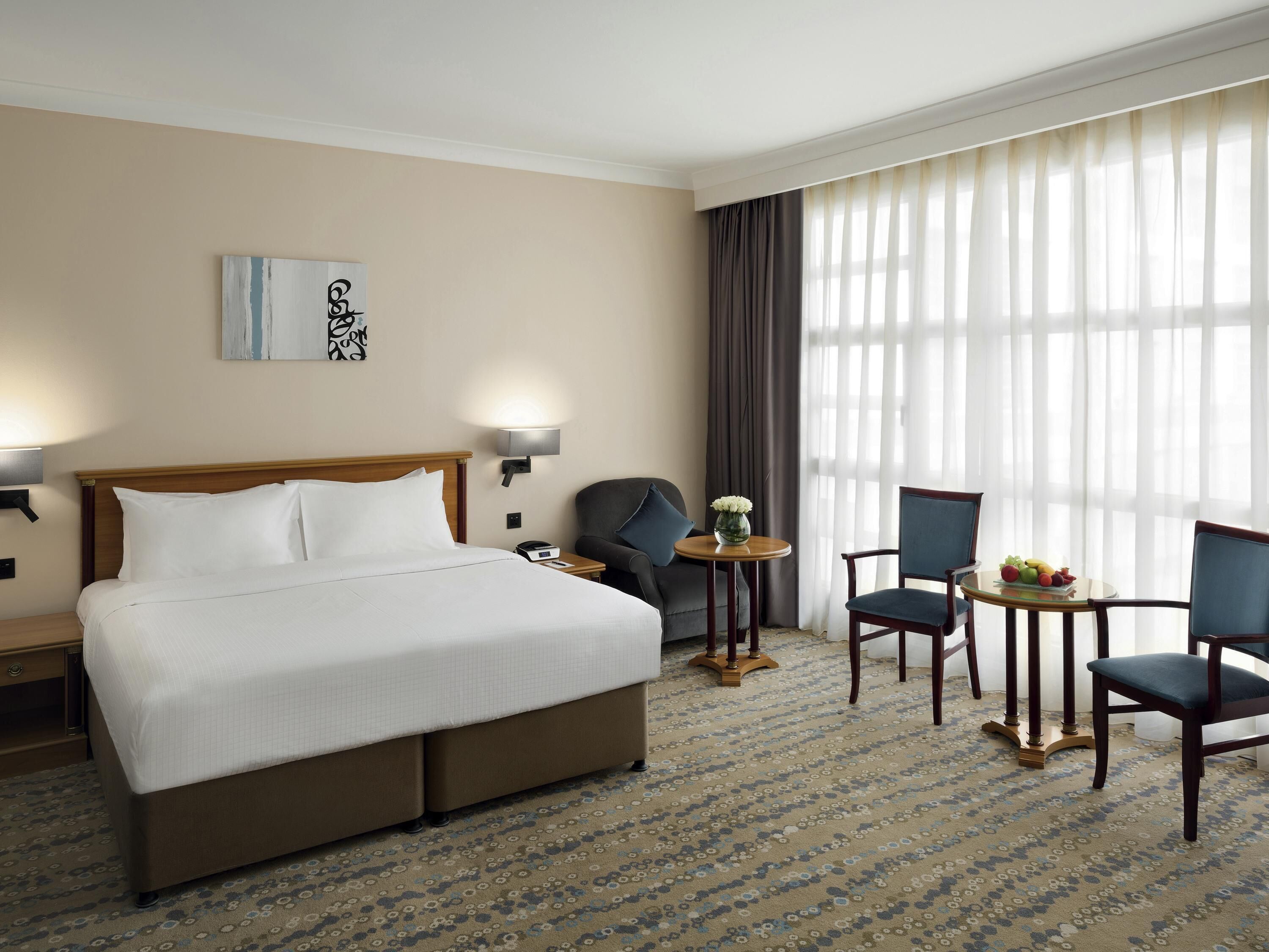 A hotel room with a large bed, two armchairs, a small table with fruit, a lamp, and a painting on the wall.