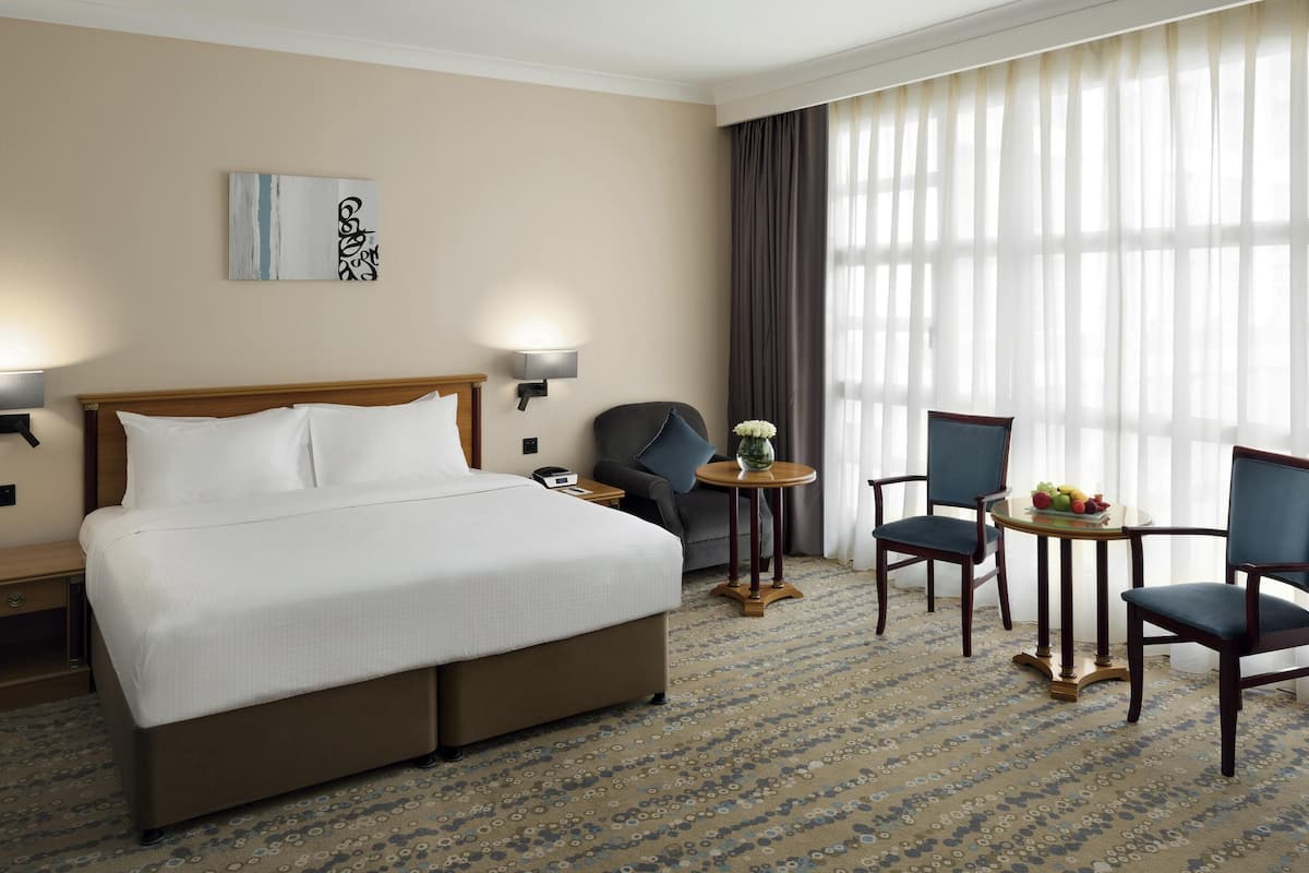 A hotel room with a large bed, two armchairs, a small table with fruit, a lamp, and a painting on the wall.
