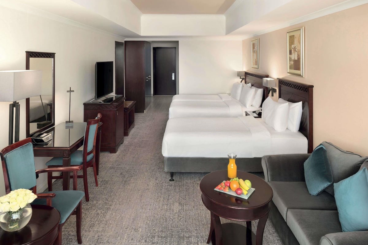 A hotel room with a bed, two bedside tables, a desk with a chair, a sofa, a small round table with fruit, and a television.