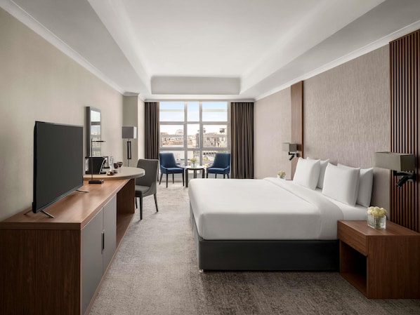 A hotel room with a bed, desk, TV, two chairs, a small table, and a city view.